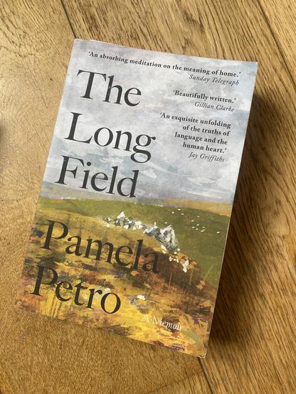The image features the cover of a book titled "The Long Field" by Pamela Petro. The cover includes various quotes praising the book, set against a background that appears to depict a landscape. The book is presented on a wooden surface.