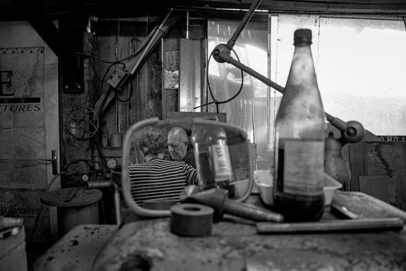 In the clutter (bottle, tools, lamp, etc.) of a DIY workshop sits a broken rear-view mirror. In the mirror we see the reflection of two men talking.