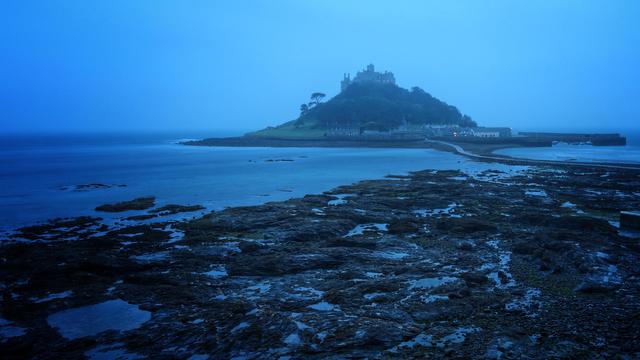 A rocky shore line at night. Just off the coast, is a small island with a castle on a hill reaching up into the sky. A causeway to the island snakes out into the sea. The picture is very blue, having been taken after sunset.