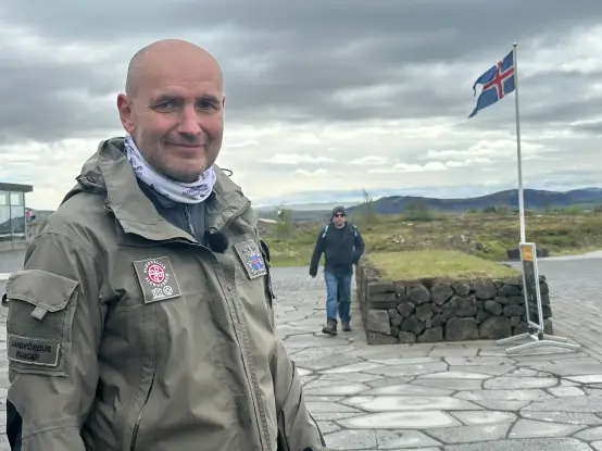 A bald man wearing a green outdoor jacket stands facing the camera. In the background is a paved surface and a flag pole with the Icelandic flag drawn. Further in the distance is the silhouette of the mountainous landscape.