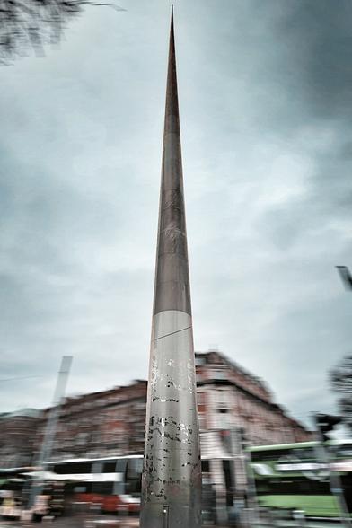 A low-angle, slightly blurred shot of the Spire of Dublin, a tall, needle-like monument, reaching towards a cloudy, overcast sky. In the background, blurred buildings and the suggestion of double-decker buses add to the urban scene. The overall impression is one of movement and the imposing height of the Spire.