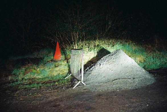 Image: Feá McCoy: Untitled, 35mm colour negative film photograph, 2024 | a photo taken at night; a bright light is shining on a mound of gravel (?) by the side of a road (perhaps); a metal crowd barrier stands to the left of the mound, and behind both we see a bright red traffic cone among grass, and other plantlife, and behind ti all, darkness• “Using analogue mediums with a particular focus on slide film and the performance of projection, McCoy’s work describes the relationship between memory, place, and grief. Navigating the past through archival family material such as photographs and home videos, McCoy creates work that is an act of remembering and reimagining, attempting to construct a fantastical past and uncover alternate versions of a real one.⁠…”