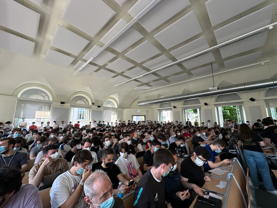 L'immagine mostra una grande sala da conferenze ben illuminata con un soffitto alto e finestre ad arco che lasciano entrare la luce naturale. La sala è piena di persone sedute a banchi, tutte indossano mascherine. La maggior parte dei partecipanti sono giovani studenti, alcuni prendono appunti o usano i loro dispositivi. . Sul lato destro della sala, una persona in piedi indossa una maglietta con la scritta "X LABS", suggerendo che potrebbe essere un organizzatore o un relatore. L'atmosfera è quella di un evento formale, probabilmente un corso, un seminario o una conferenza.