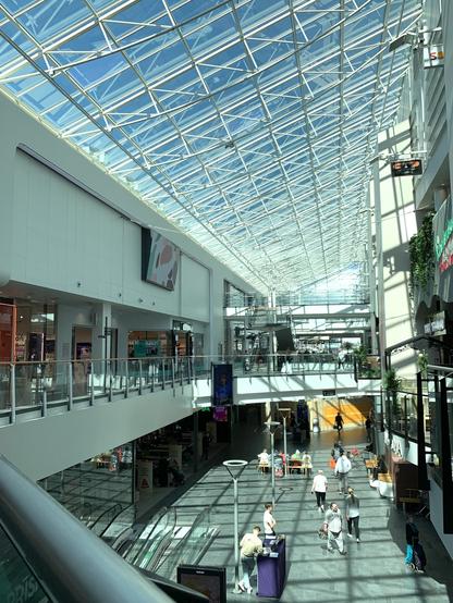 A modern shopping mall interior with a glass roof, allowing plenty of natural light. The space features multiple levels, escalators, shops, and people walking through the area. Shadows and reflections create visual interest on the floor.