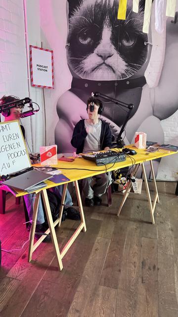 Das Foto zeigt zwei junge Personen, die an einem gelben Tisch sitzen und einen Podcast aufnehmen. Sie tragen große Kopfhörer und sprechen in Mikrofone, die an Mikrofonarmen befestigt sind. Auf dem Tisch liegen verschiedene Gegenstände, darunter ein Audio-Mischpult, Notizbücher und Flyer. Im Hintergrund befindet sich ein großes Wandbild einer Katze mit finsterem Blick, die wie ein Astronaut dargestellt ist. Links neben dem Tisch steht ein Whiteboard mit der handgeschriebenen Aufforderung: "Nehmt euren eigenen Podcast auf." An der Wand hängt ein Schild mit der Aufschrift „Podcast Station“. Der Raum ist in pink-violetten Lichttönen beleuchtet, der Boden besteht aus dunklem Holz, und insgesamt herrscht eine kreative, entspannte Atmosphäre.