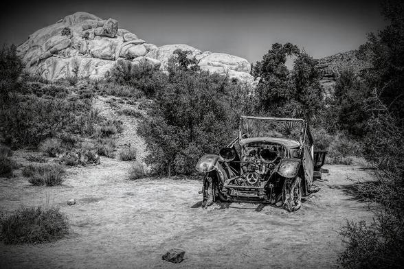 An old, rusted car sits abandoned on the Wall Street Mill Trail in Joshua Tree National Park, surrounded by desert landscape and rugged beauty.