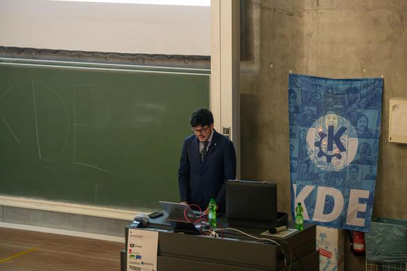 A photo from Akademy 2024 shows a speaker addressing the audience on stage.