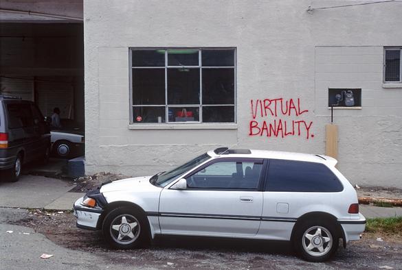 A small white park is parked in front of a white mechanic's garage window, with the words "VIRTUAL BANALITY." spray painted in red on the wall by the bottom right corner of the window.