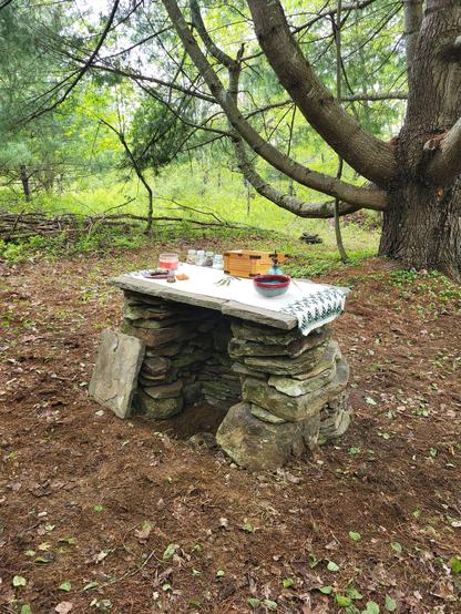 A dry stone altar. It is dressed with an altar cloth and ritual implements, and an open chest awaiting offerings. The hearthstone has not been placed yet in this photo, as the photo was taken before the consecration. It has since been consecrated and the hearthstone placed. Beyond the altar is a very large, very old white pine tree we call Great Great Grandfather Pine. He is a wonderful listener.