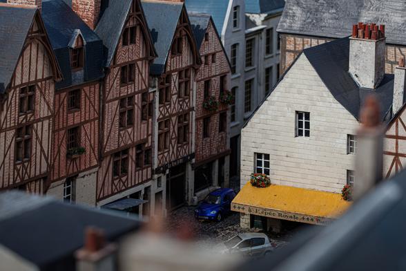 Aerial view of a miniature model depicting European-style buildings, with a focus on a white building with a yellow awning labeled “L'épée Royale” and miniature cars parked nearby.