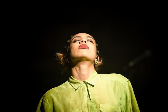 Eine Sängerin mit gelber Bluse vor dunklem Hintergrund. Sie richtet ihren Kopf mit geschlossenen Augen nach oben.
