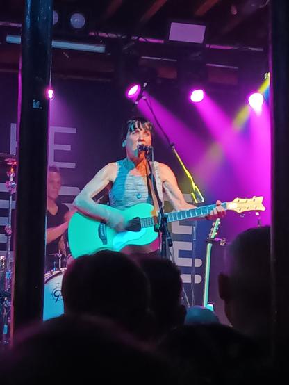 Kristin Hersh performing with her band, Throwing Muses. She is a small, white woman with back hair, standing at a microphone and playing a turquoise guitar.
