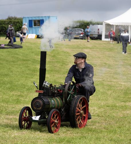 A bearded man sitting on a small steam-powered green traction engine, which has smoke coming from the chimney