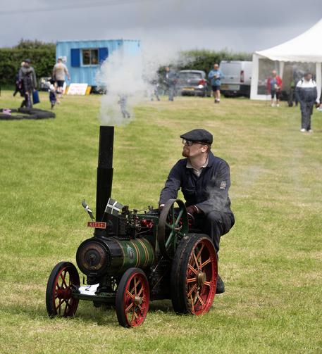 A bearded man sitting on a small steam-powered green traction engine, which has smoke coming from the chimney