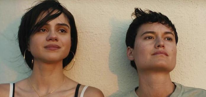 Sasha Calle and Lio Mehiel play the adult siblings in In the Summers. Here they are leaning against a stucco wall with the sun on their faces. Both are dark haired, staring into the distance.