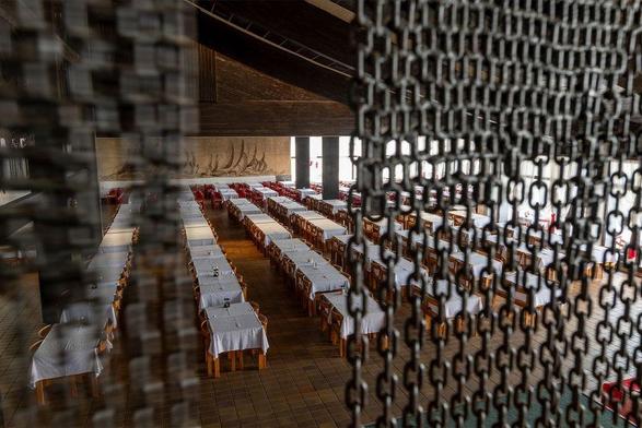 Image: Barbara Knežević: Gvozdene Kapije (The Iron Gates), 2024, production still. Photography by Louis Haugh | close to the camera, chains hang down from above, parted towards the middle, and partially hiding a view of a very large refectory; maybe 50 tables are lined up in rows, each with a white tablecloth and four chairs and what may be some condiments; there are supporting columns and what may be signs of a sloping ceiling; a far wall has a large hanging or mural, which appears to be depicting sailboats at sea • “The Iron Gates is an exhibition of new sculpture and film work by Barbara Knežević commissioned by Solstice Arts Centre and supported by the Arts Council of Ireland. The exhibition is a series of sculptural objects and a single-channel film that interprets the history, stories, materiality, and the development of technology in the Iron Gates (Gvozdena kapije) region, a deep scenic gorge at Đerdap on the Dunav (Danube) River on the border of Serbia and Romania. It is…”