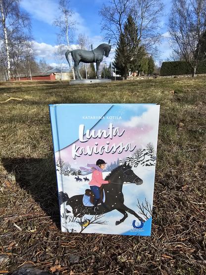 Kuvassa Lunta kavioissa -kirja ja taustalla näkyy hevospatsas.