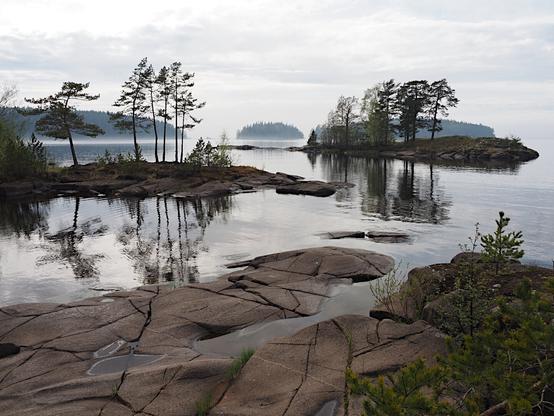 A view of a set of islands by the lake shore, large and small. Pine trees on the islands reflect in the waters. There are bigger islands in the background, visible through the mist over the cold waters. The sky is overcast with grey clouds.