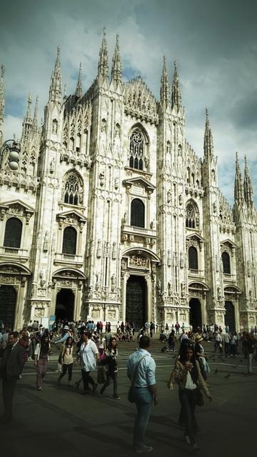 A low-angle shot captures the intricate, white marble facade of the Milan Cathedral under a dramatic, cloudy sky. Numerous detailed spires and statues adorn the building's exterior. In the foreground, a diverse crowd of people is walking across the piazza, some looking towards the cathedral, others moving through the square. The scene is bustling with activity, highlighting the grandeur of the cathedral against the daily life of the city.