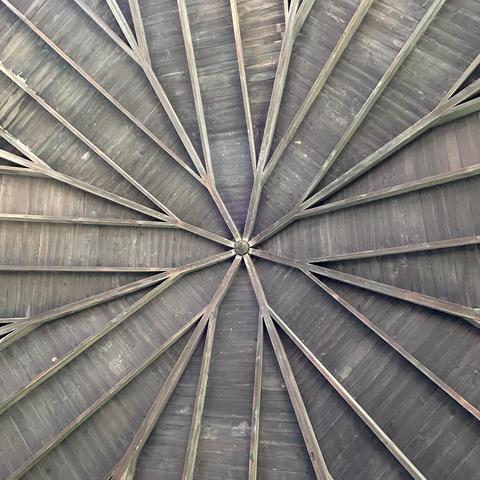View looking up at a wooden ceiling, the beams coming together at a central point and radiating outward in an eight-fold design kind of like a flower.