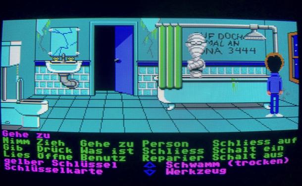 Screenshot on a CRT monitor showing the bathroom in Maniac Mansion. A broken mirror and sink next to a toilet. Also a  mummy sitting in a bathtub. The player is currently controlling Dave. At the bottom an array of action verbs and the inventory can be seen. The game language is German.