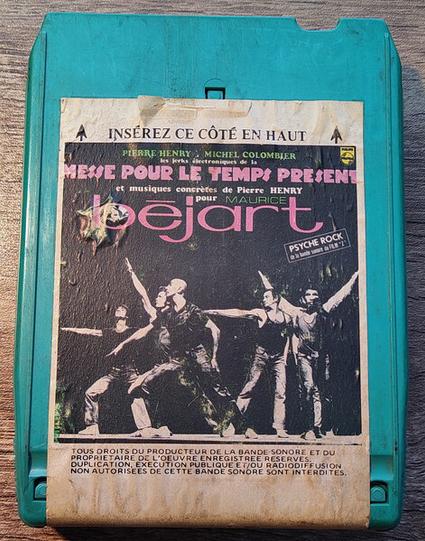 A photo of a vintage light blue plastic 8-track cartridge tape by Pierre Henry and Michel Colombier entitled "Les Jerks Électroniques De La Messe Pour Le Temps Présent Et Musiques Concrètes Pour Maurice Béjart" on a woodgrain background. Inset black and white photo shows 6 men on a stage posing with arms outstretched. All printed text is in French.