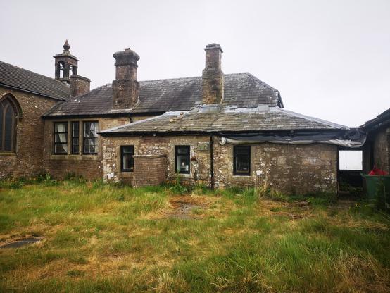 An old single storey schoolhouse with, on the left, a  small church attached. In front of the school is an extension with a galley kitchen inside. It's Grey and just about to rain.