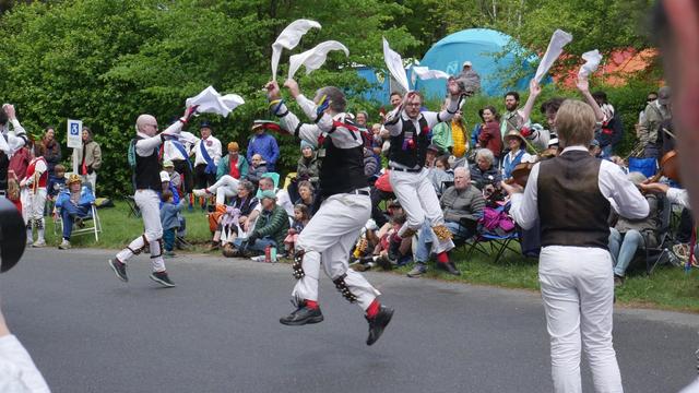 Three morris dancers with white shirts and trousers and black vests, decorated with multicolor ribbons, dancing on a paved surface. All are in motion, feet off the ground and arms raised. Two musicians play fiddles on the right. Behind them is an audience including a number of other morris dancers.