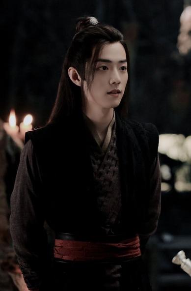 Wei Wuxian in the Burial Mounds during Lan Wangji's visit. They are in the cave, and there are lit candles in the background. Wei Wuxian is wearing dark robes, black over a more faded black belted with red over more black, and at his neck is visible a more earth-tone underlayer. He is facing Lan Wangji, whose presence is indicated by the very top bit of Bichen's hilt visible in the lower right of the image.