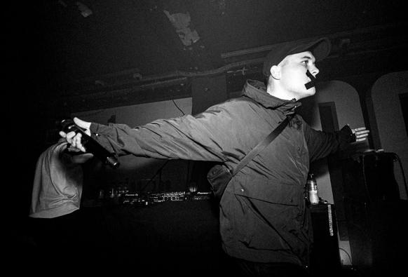 Black and white image of Mo Chara from Kneecap on stage.  He's got black scotch tape across his mouth.