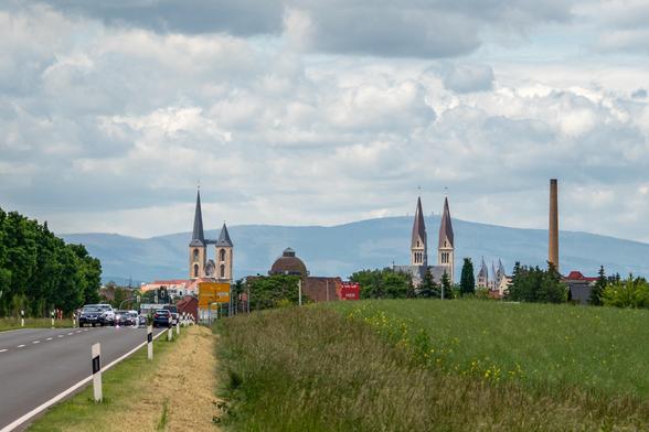 Blick auf eine Skyline, im Hintergrund ist der Brocken zu sehen, im Vordergrund rechter Hand eine Straße, in der Mitte und linker Hand ein grünes Feld, am Horizont die Silhouette einer Stadt (Halberstadt) mit Martinikirche, Dom und einem Schornstein