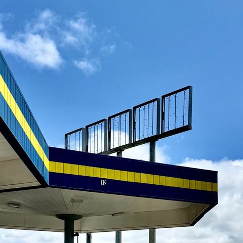 Blue and yellow striped gas station canopy and empty sign holder against a blue sky with fluffy white clouds.