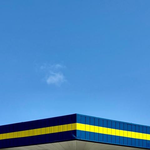 Another angle of the blue and yellow striped gas station canopy, this time from the center so both sides are going down forming a sort of triangle even though the canopy itself was more of a hexagon. Above is a bright blue sky with a small wispy cloud.