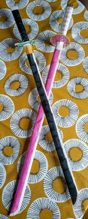 Two Japanese katana (?) crafted from paper. One is pink with orange accents, the other black with green and gold.