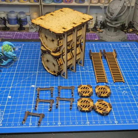 A detailed assembly of the TT Combat Sector 4 Storage tanks is displayed on a blue cutting mat marked with a grid. The main structure consists of two large, cylindrical tanks stacked vertically, supported by a wooden frame with intricate detailing. Surrounding the tanks are various components, including two sets of ladders, several hazard markers labeled "DANGER," and additional structural pieces, all crafted from a light wood material. In the background, a miniature figure and a globe stand are visible, adding context to the tabletop gaming environment.