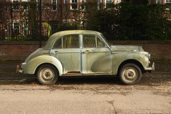 A vintage car in a light teal colour but covered in green algae and dirt. The car is rusted away in places. It is parked at the side of the road with a low brick wall, vegetation and buildings behind.