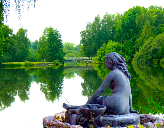 Fotomontage von 2 Fotos
Eine weibliche Skulptur sitzt an einem See und blickt auf die Bäume, die sich im See spiegeln. Weiter im Hintergrund gibt es eine alte Holzbrücke, die über den See führt.