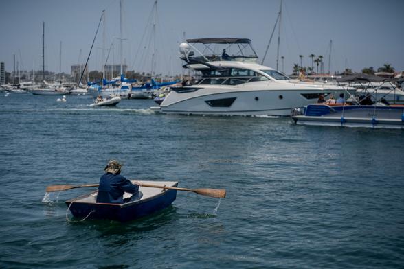 A bustling marina scene on a calm day. In the foreground, a person in a blue shirt and camouflage hat rows a small blue wooden boat using wooden oars. The rower is facing away from the camera, moving towards a busy harbor filled with various boats.

In the middle ground, a large white luxury yacht is prominently visible, its sleek design contrasting with the smaller sailboats around it. The yacht appears to be in motion, creating a small wake.

The background shows numerous sailboats with tall masts, some with blue covers. Palm trees are visible on the distant shoreline, suggesting a tropical or subtropical location. The sky is clear and blue, adding to the serene atmosphere of the scene.