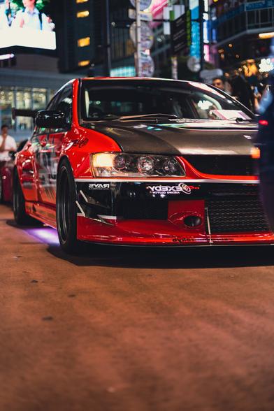 photo of a Mitsubishi Lancer Evolution VII tuning car from the front, with its headlights in focus. It's night on y lively and well illuminated street.
