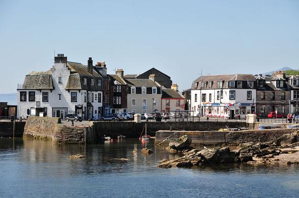 Millport. The image shows a view from the sea of a stone harbour with some small boats in it. Beyond the harbour are mainly two-storey buildings forming shops and other commercial buildings. The sky is blue and the sea is reflecting it.