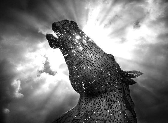 Image shows one of the pair of Kelpies , horse head water spirits, works of art just outside of Falkirk 
The horse stands in front of an afternoon sun, with sun beams shooting left and right from the horses head