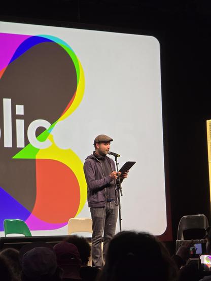 Der Autor Marc-Uwe Kling, mit Pullover, Jeans und Cap bekleidet, steht an einem Mikrofon mit einem Tablet in der Hand.