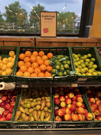 A display of fresh fruits in a grocery store, featuring oranges, lemons, limes, pears, and apples. There's a sign that reads "Gratis frugt til børn," indicating free fruit for children while parents shop.