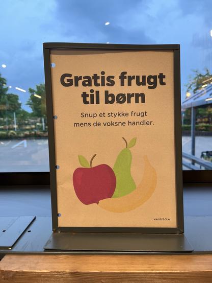 A sign displaying a message in Danish offering free fruit for children while adults shop. The background includes illustrations of an apple, a pear, and a banana.