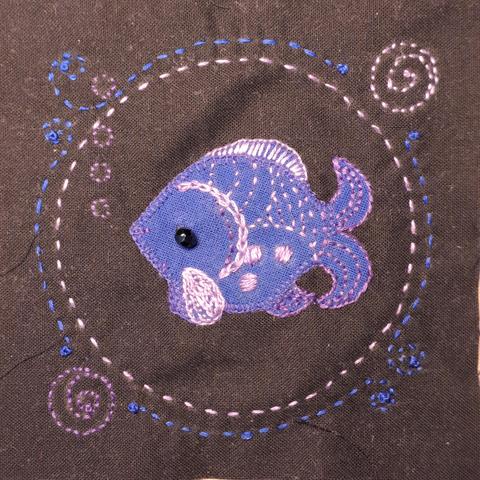 Auf einem schwarzen Quadrat Baumwollstoff wurde ein Fischchen aus blauem Baumwollstoff aufgestickt.