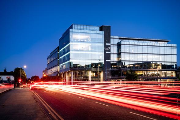 A modern glass office building at dusk in Cork, Ireland, with illuminated interiors and the names "Johnson Controls" and "ARUP" visible on the facade; red and white light trails from passing vehicles streak across the road in the foreground, under a deep blue sky.