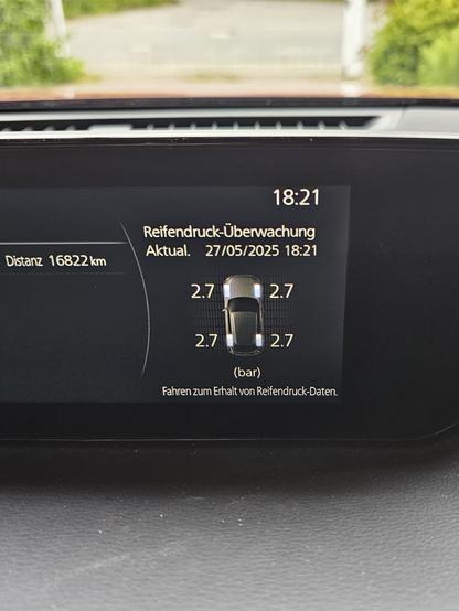Reifendruckanzeige eines Fahrzeug-Displays mit den einheitlichen Werten von 2,7 Bar je Reifen. Im Hintergrund eine Straße mit grünen Büschen.