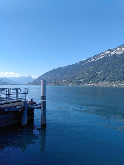 Ein Steg, blaues Wasser und rund darum Berge. Blauer Himmel.