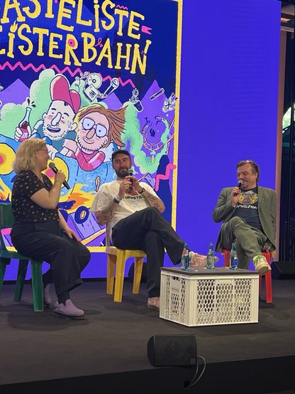 Auf der Bühne sitzen Maria Lorenz-Bokelberg, Markus Herrmann und Nilz Bokelberg und reden über Biopics, Film usw. Im Hintergrund sieht man eine bunte Graphik, die die drei Protagonisten in einer Geisterbahn zeigt...