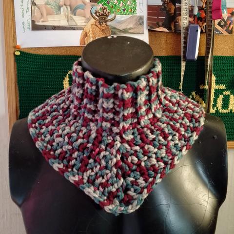 Cuello tejido a crochet con lana matizada en tonos burdeos, celeste y beige, exhibido en un busto negro frente a un tablero de corcho.
